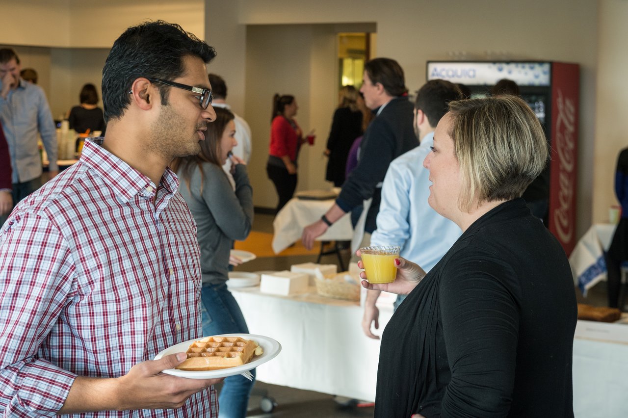 Two people converse at a breakfast event, one holding a plate with waffles and the other holding orange juice.