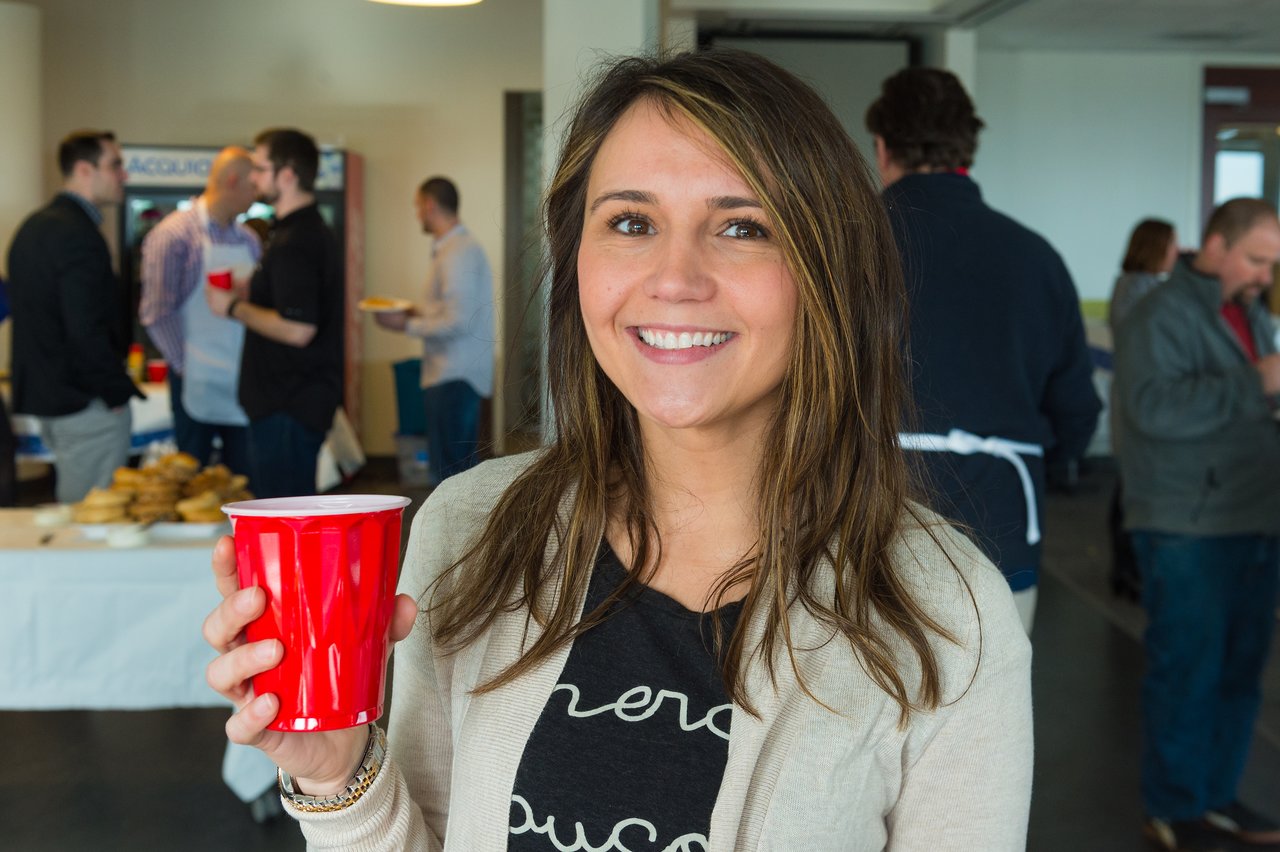 A smiling woman holds a red cup at a networking event, with people chatting and food in the background.
