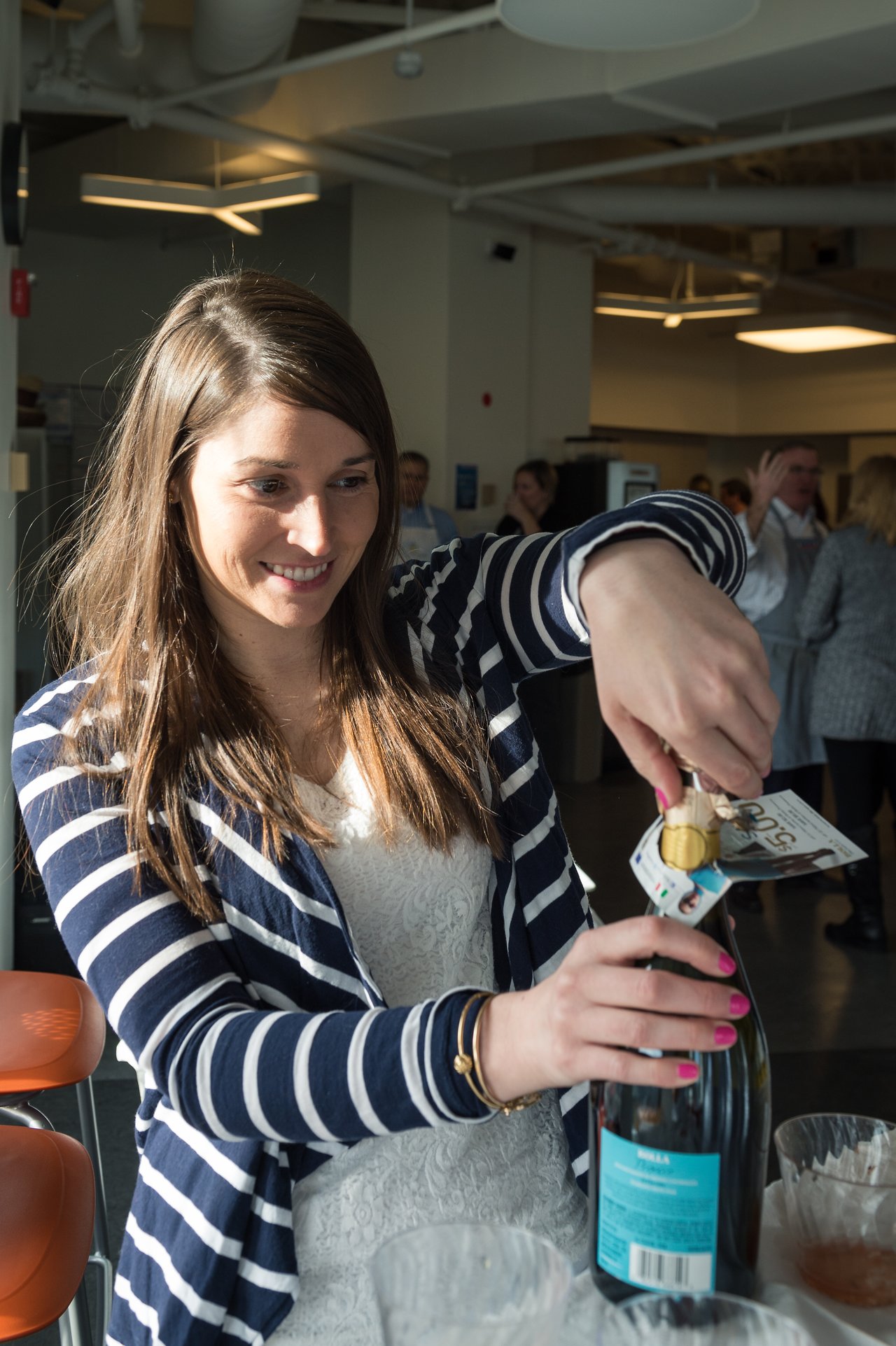 A woman in a striped sweater opens a bottle of sparkling wine at an indoor event.