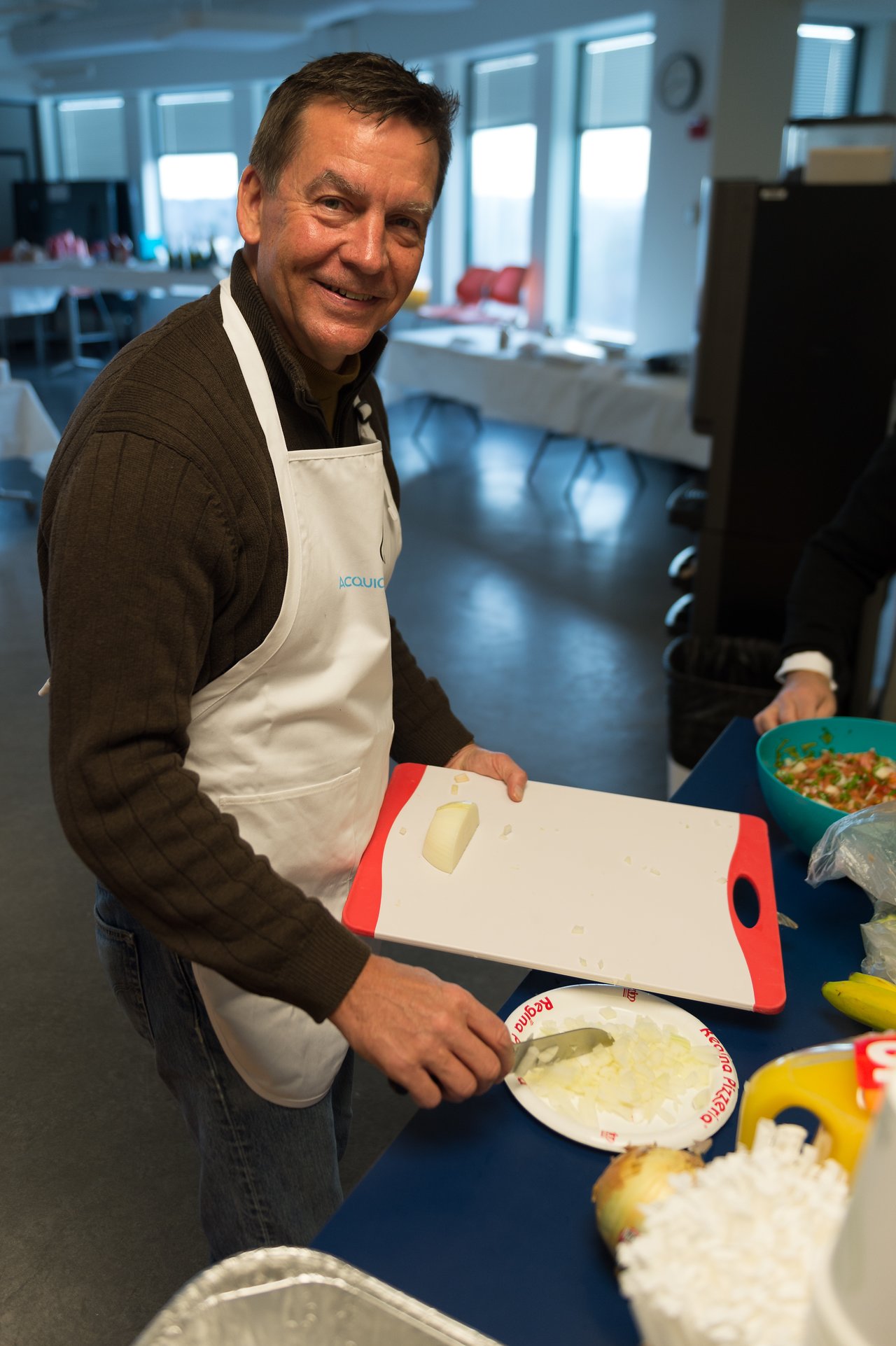 A man wearing an apron smiles while chopping onions on a cutting board at a food preparation event.