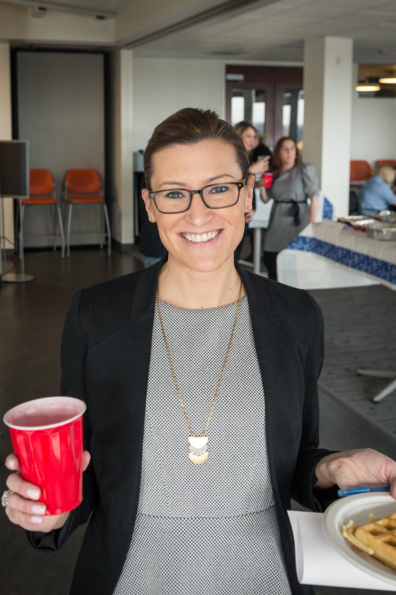 A smiling person in business attire holds a red cup and a plate of food at a networking event.