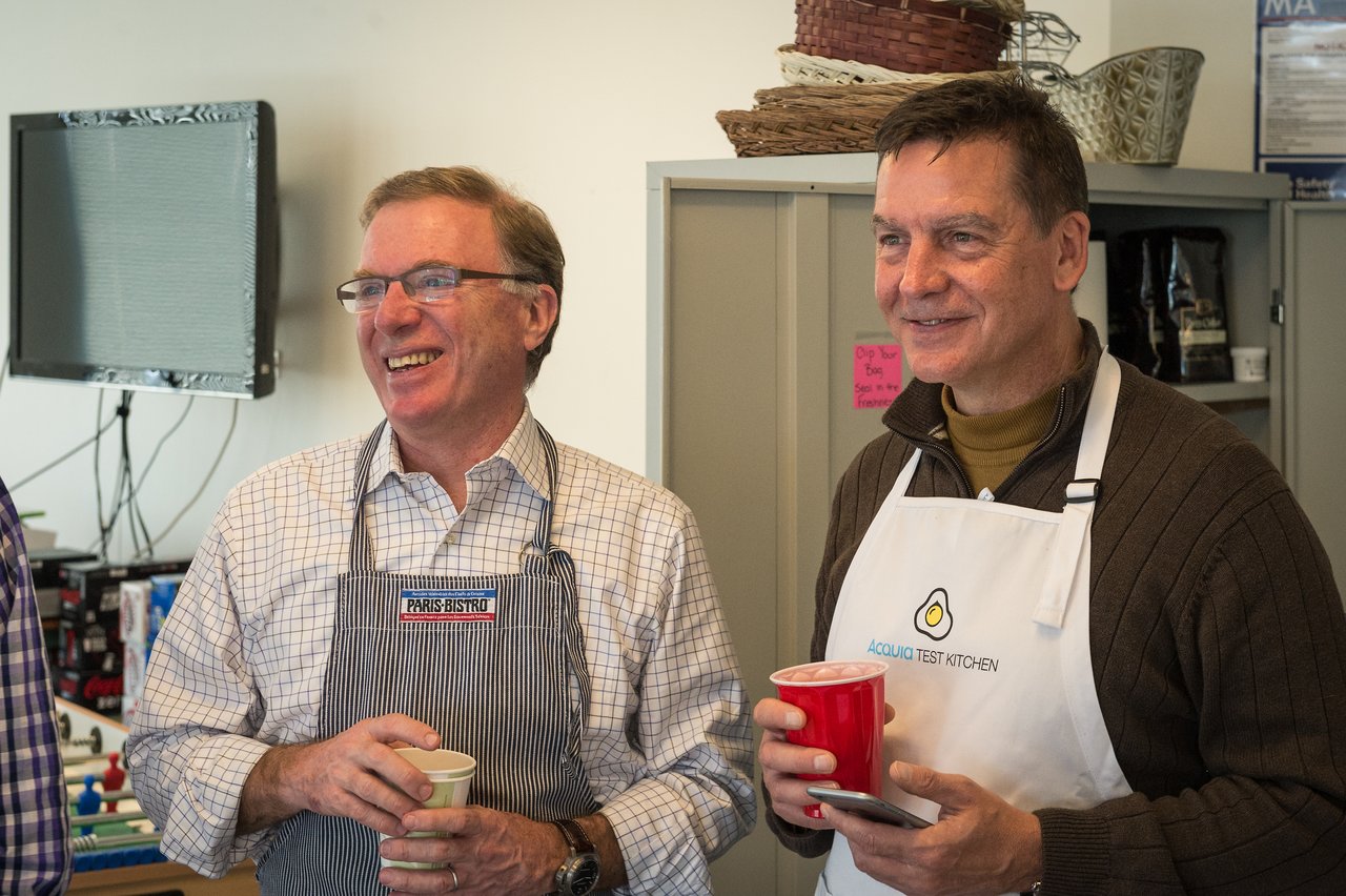 Two men wearing aprons stand together, smiling and holding drinks at an Acquia event.