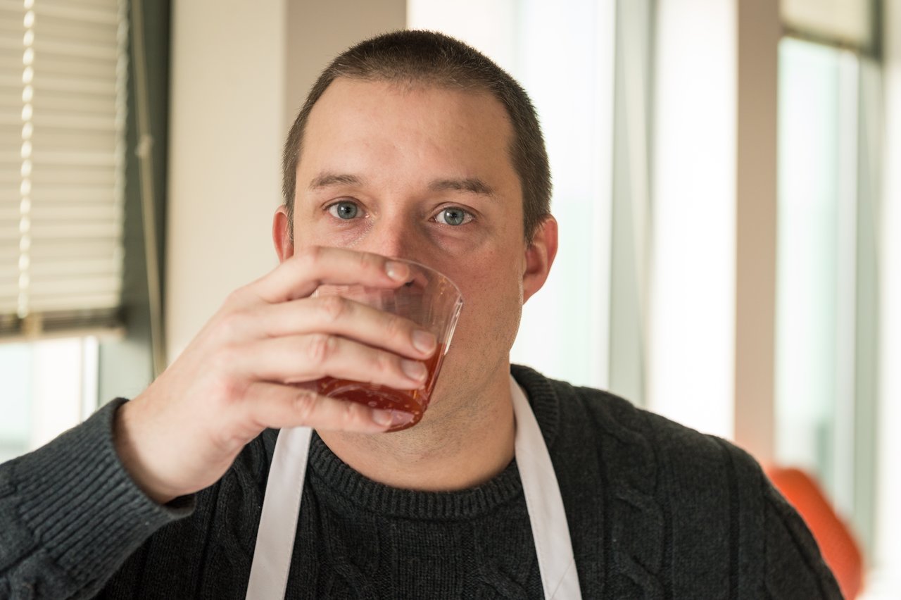 A man wearing a dark sweater and white apron drinks from a glass with a reddish liquid.