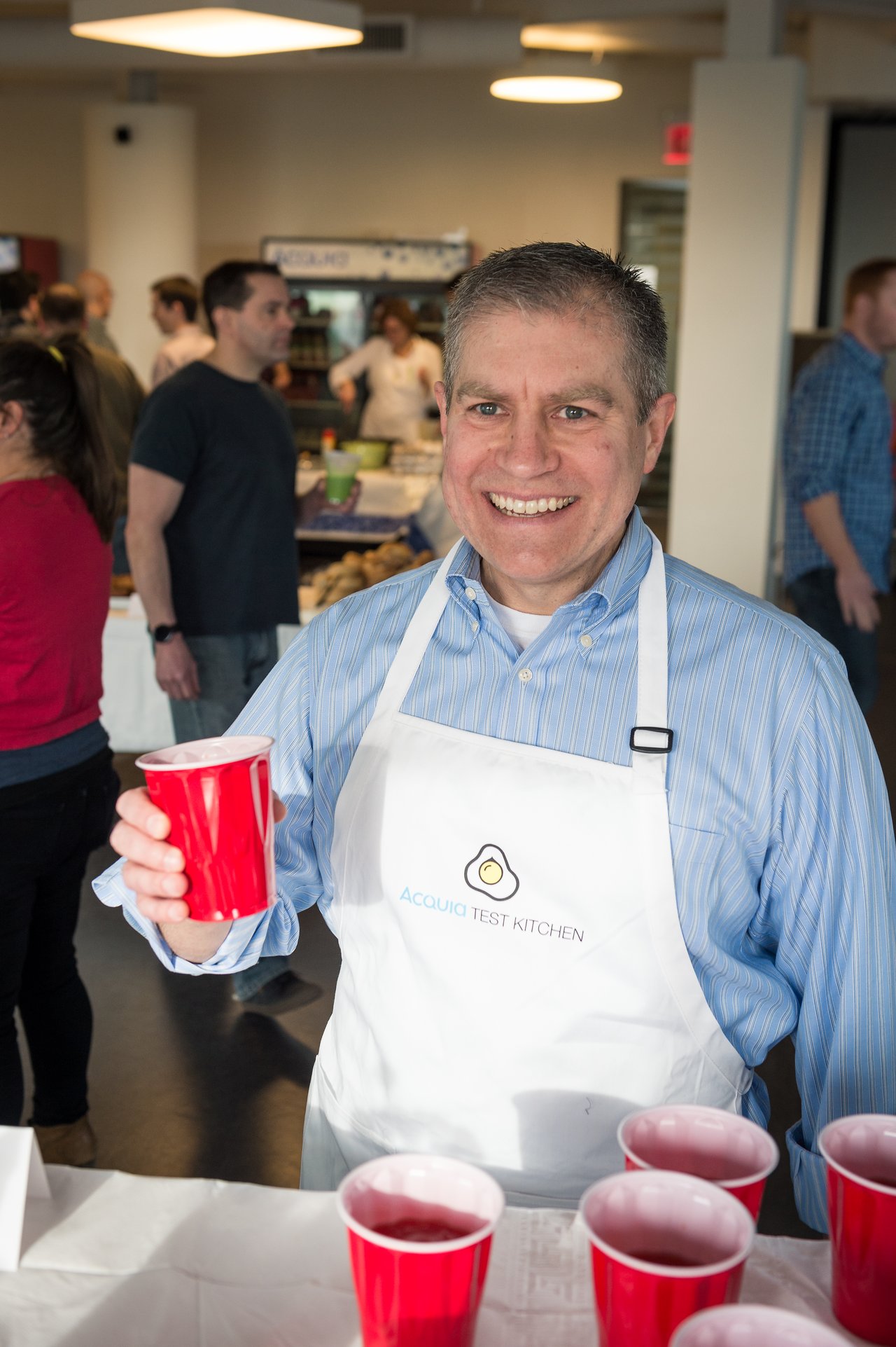 A smiling man in an Acquia Test Kitchen apron holds a red cup at a breakfast event.