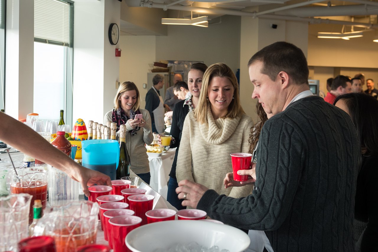 People gather around a table with drinks, chatting and serving beverages at a casual indoor event.