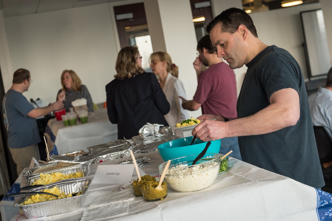 A man serves himself food from a buffet table while others converse in the background at a breakfast event.