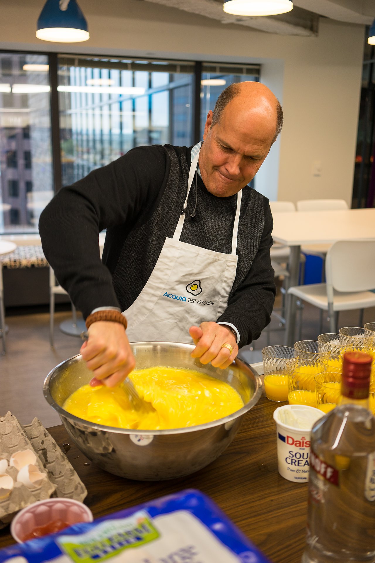 A person wearing an Acquia apron vigorously whisks eggs in a large bowl at a breakfast event.