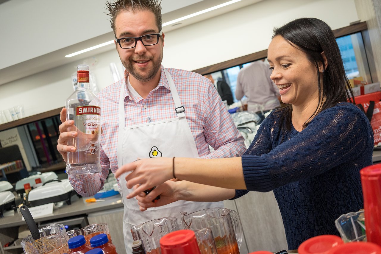 A man in an apron holds a vodka bottle while a woman smiles and prepares drinks at an event.