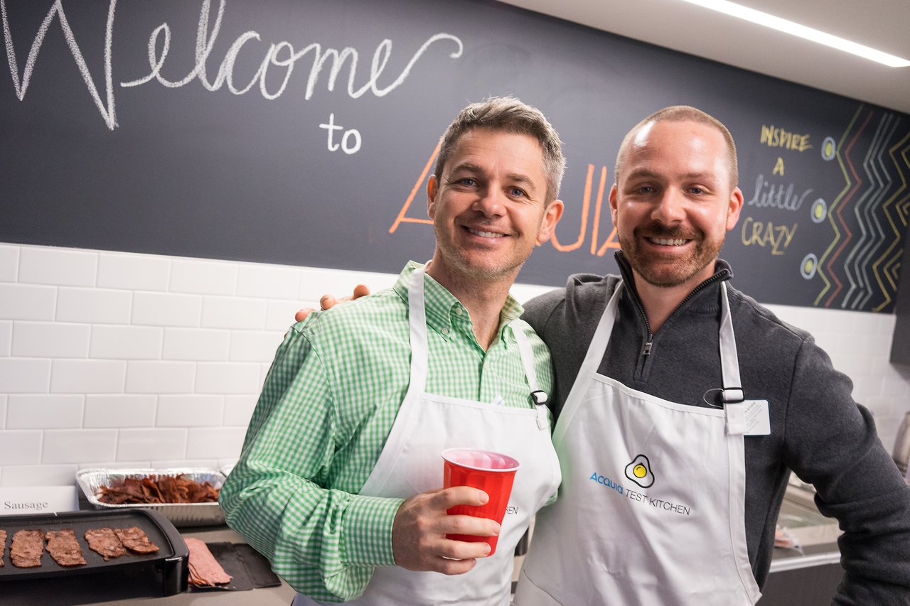 Two men wearing aprons smile and pose together at an Acquia event, with food cooking in the background.