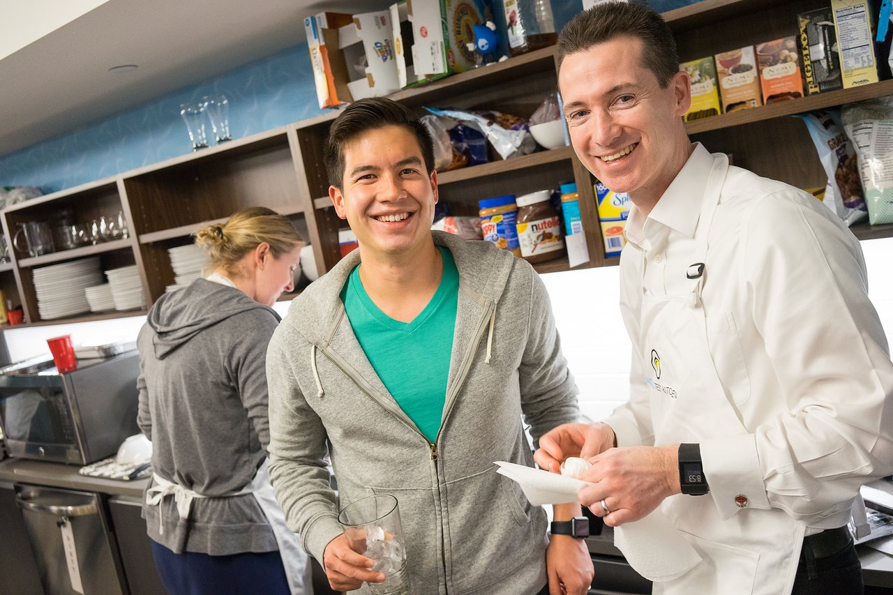 Two men smiling in a kitchen, one wearing an apron and holding a napkin, while another holds a glass.