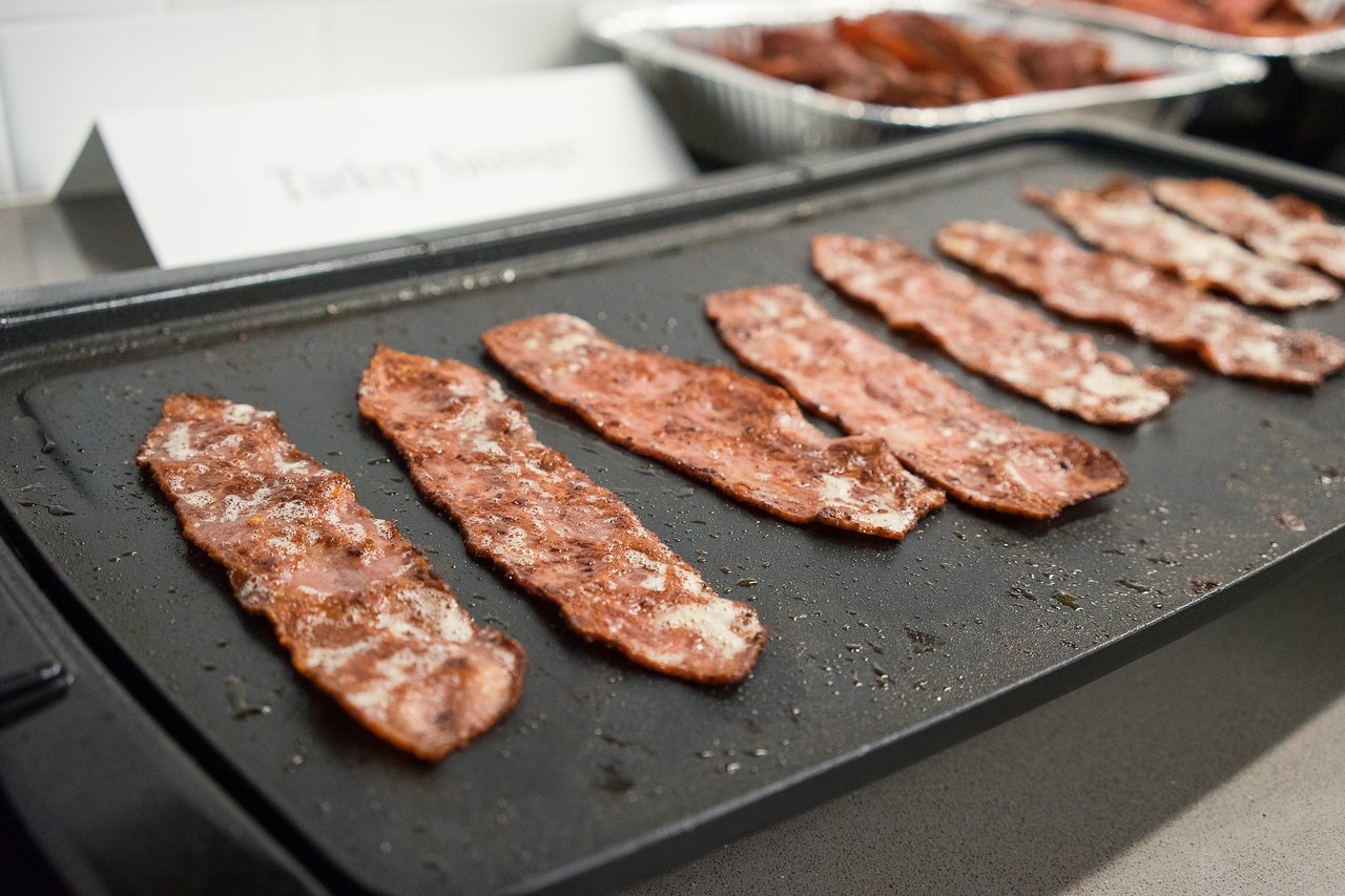 Strips of turkey bacon cooking on a griddle, with trays of cooked bacon in the background.