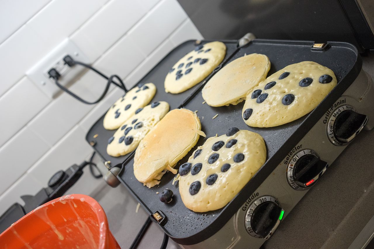 Blueberry pancakes cooking on a griddle, with one partially flipped.