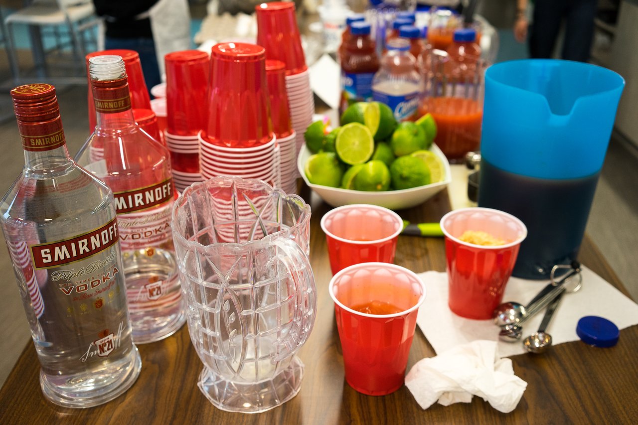 A table with vodka bottles, red cups, limes, juice, and a blue pitcher set up for drinks.