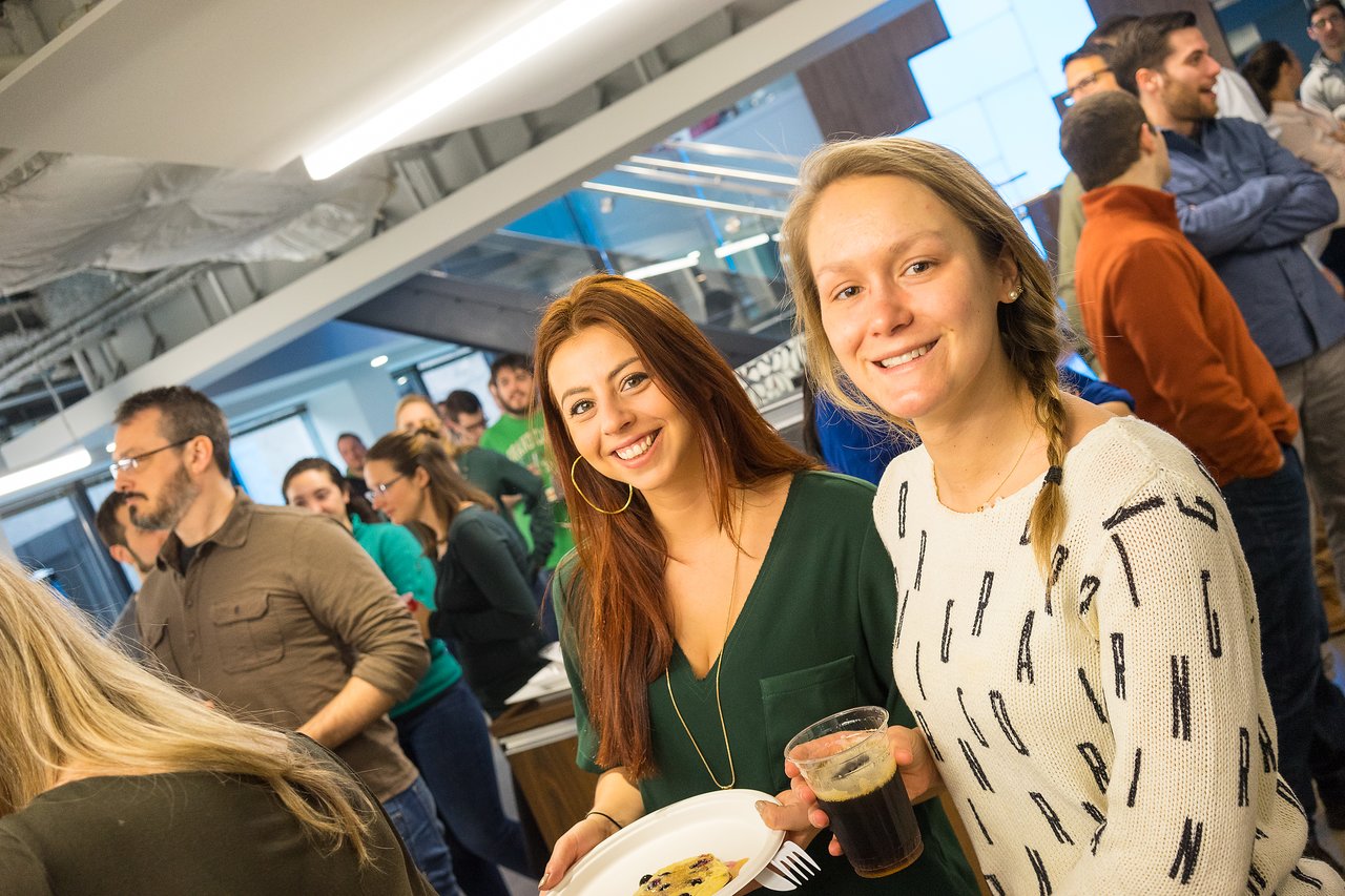 Two smiling women at a networking event, holding food and drinks, with a crowd socializing in the background.