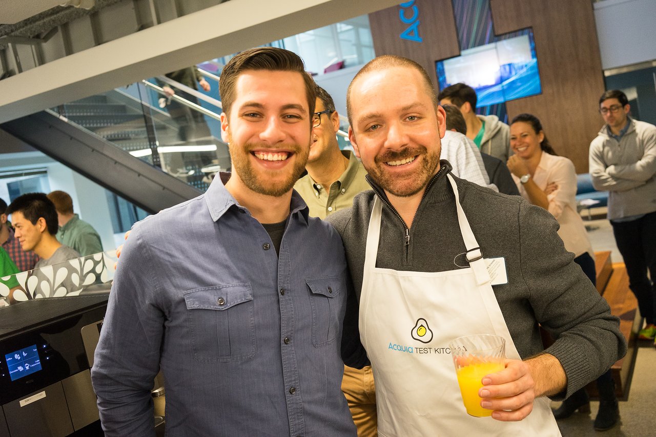 Two smiling men pose together at an Acquia event, one wearing an apron and holding a drink.