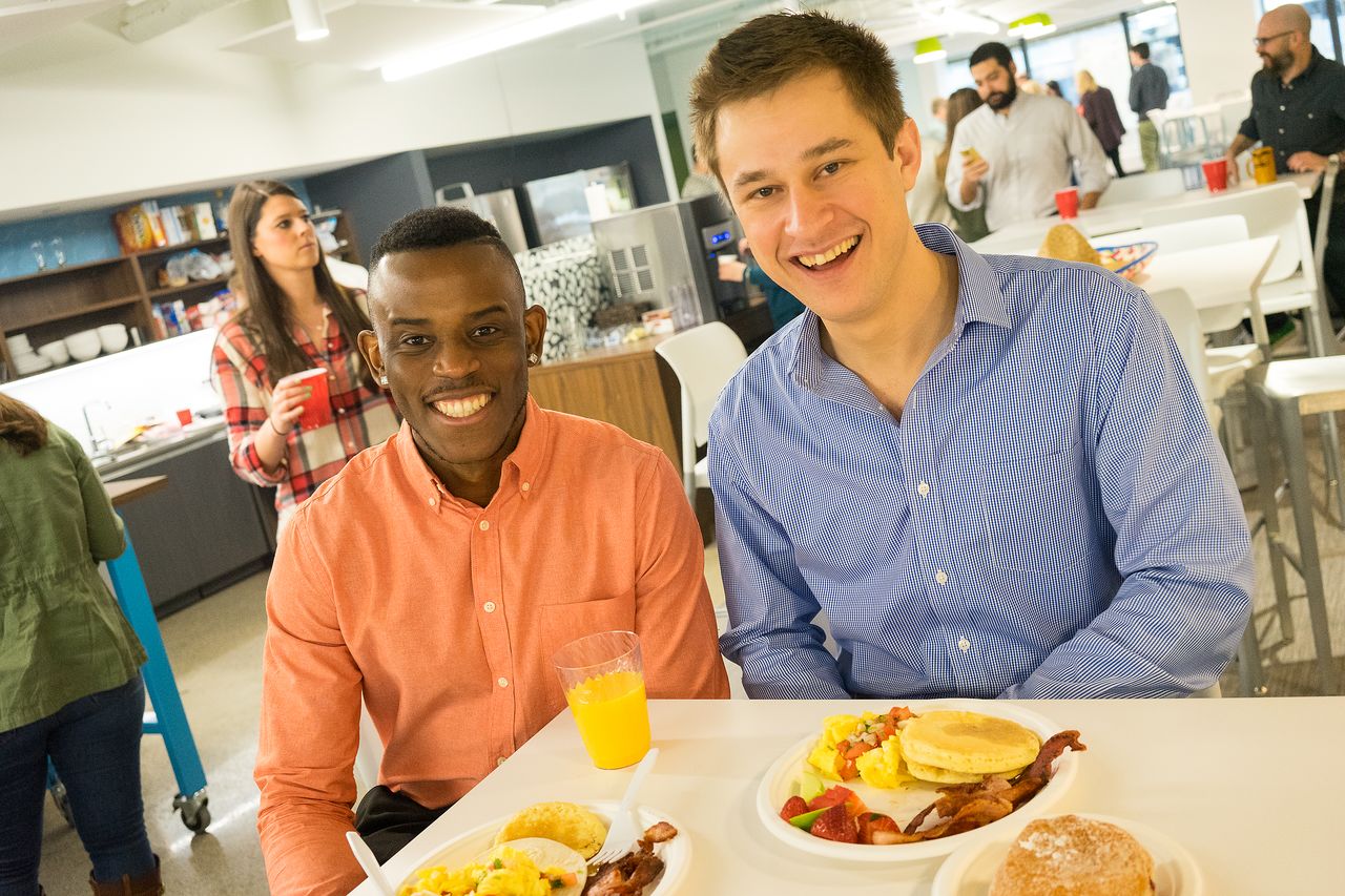 Two smiling men sit at a table with plates of breakfast food during a casual gathering in a shared space.