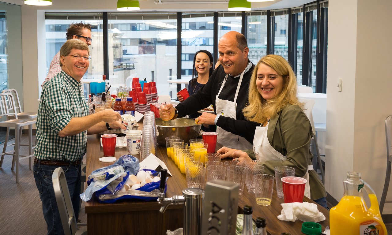 A group of people prepares and serves breakfast, smiling and interacting in a bright office setting.