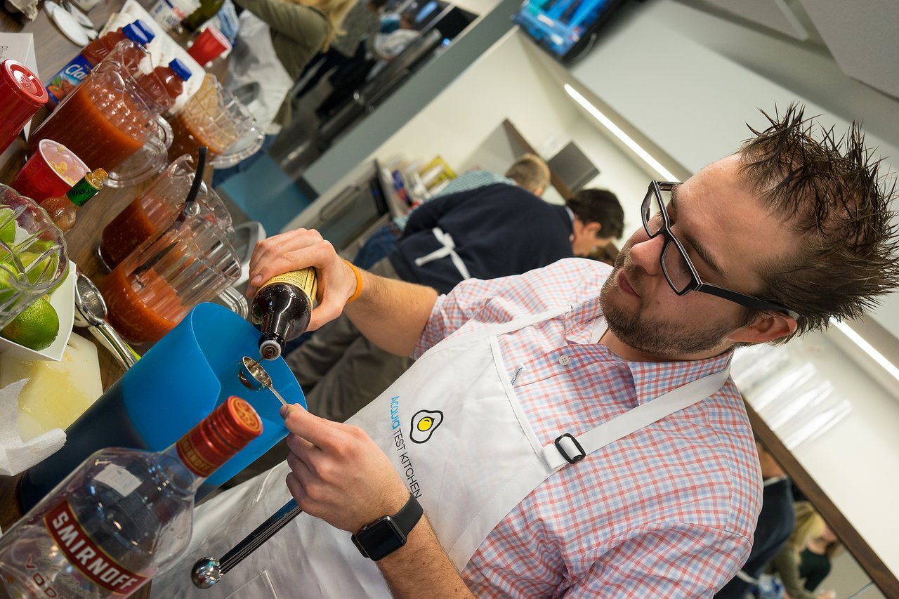A man wearing an apron pours sauce into a spoon while preparing a drink at a kitchen event.