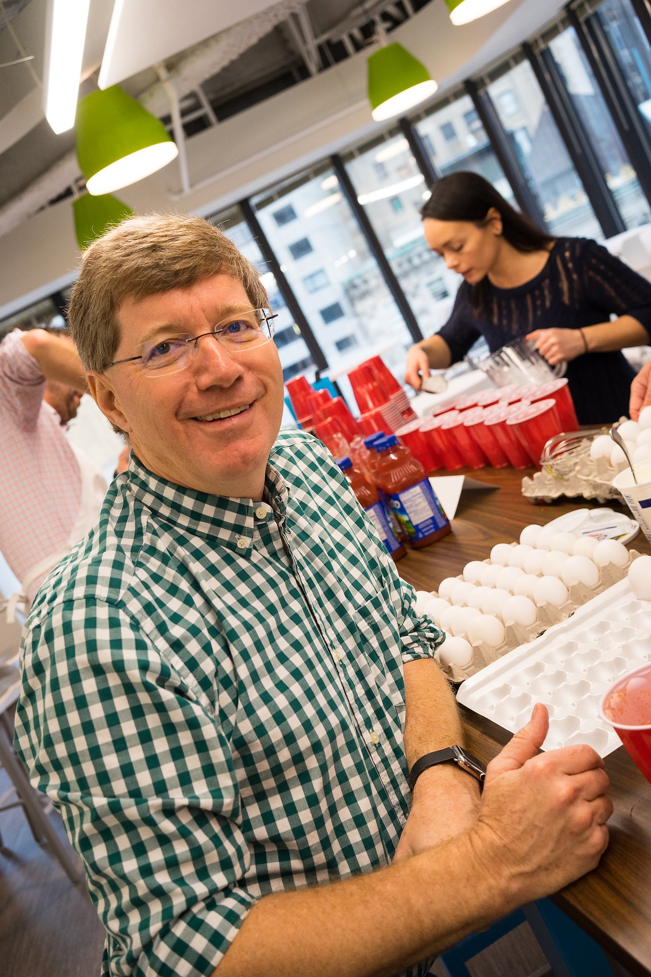 A smiling man in a checkered shirt stands near a table with eggs and drinks at a gathering.