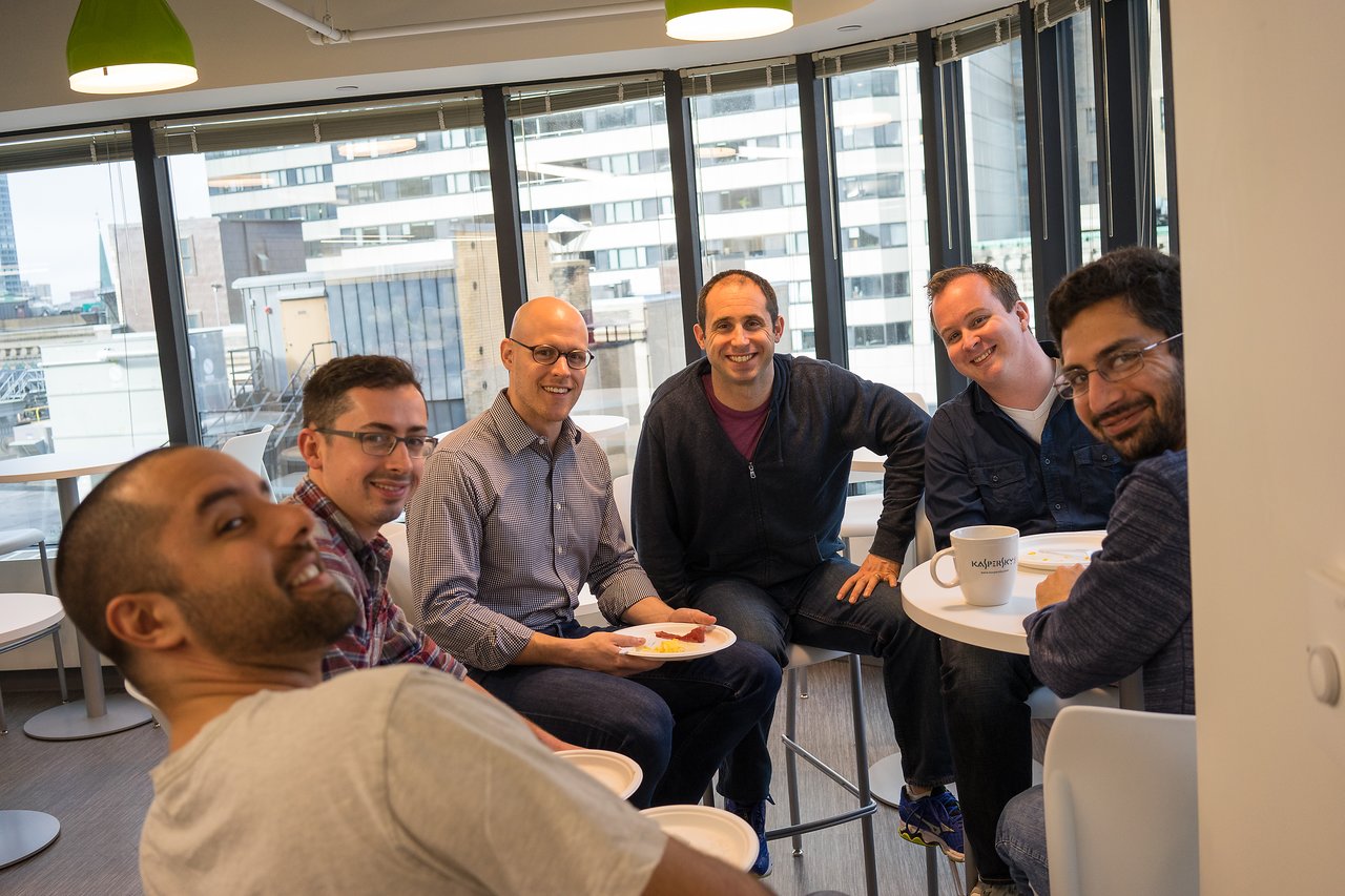 A group of six people sitting at a table, smiling and enjoying breakfast in a bright office space.