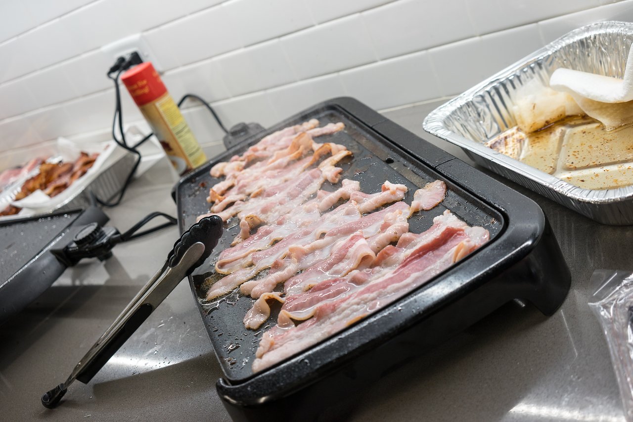 Bacon cooking on an electric griddle with tongs resting nearby at a breakfast event.