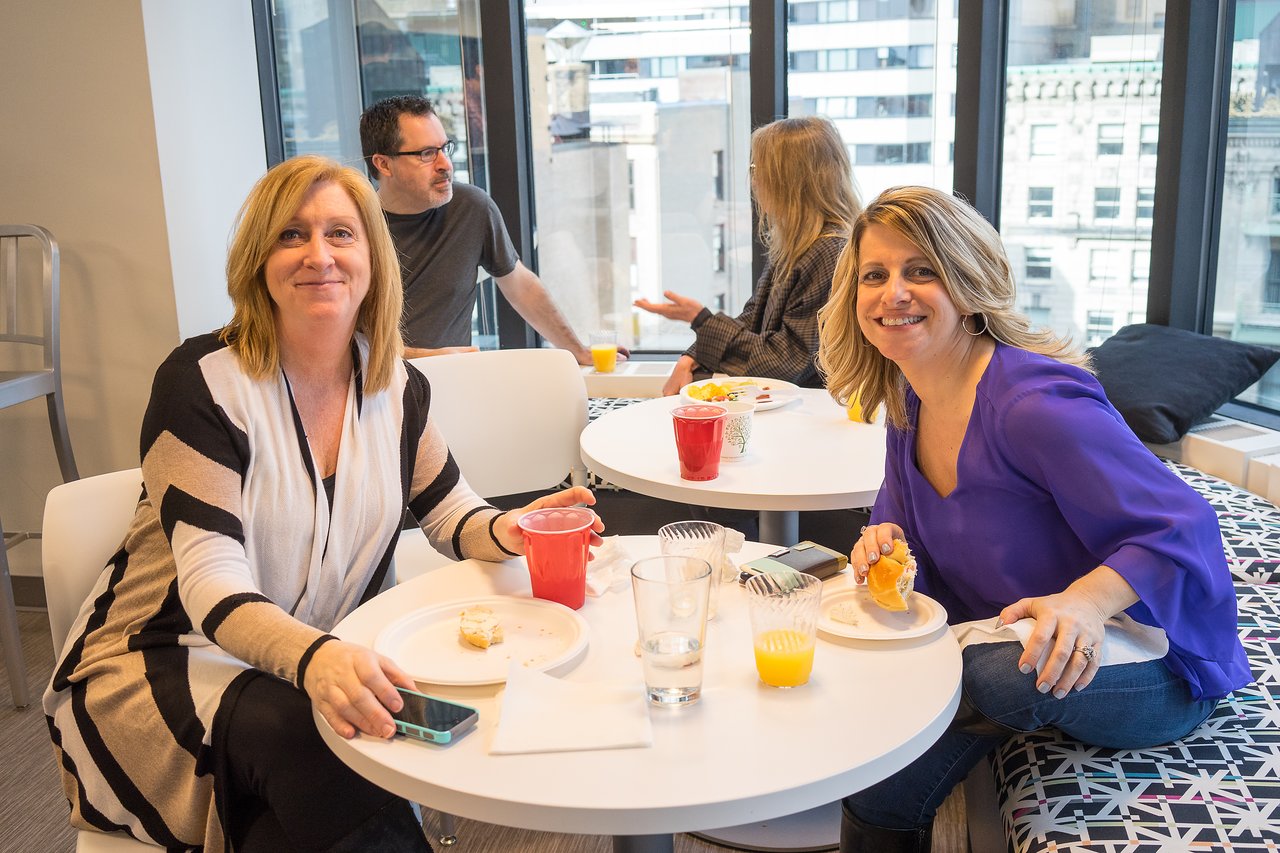 Two women sit at a table, smiling and eating breakfast, while two others talk in the background.