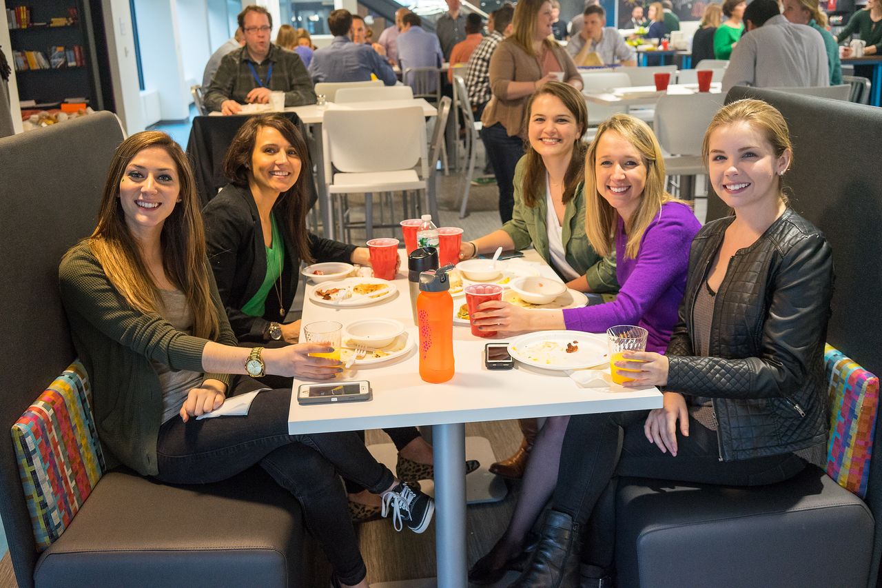 A group of five women sitting at a table, smiling and enjoying food and drinks at a social event.