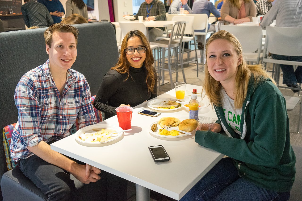 Three people sitting at a table, smiling and eating breakfast at a casual event.