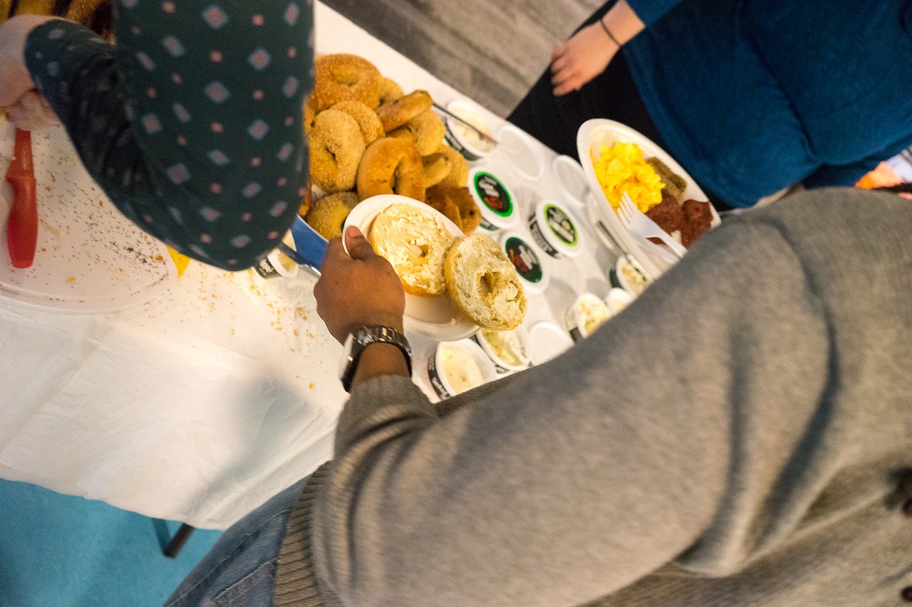 People serving themselves bagels and spreads at a breakfast buffet table.