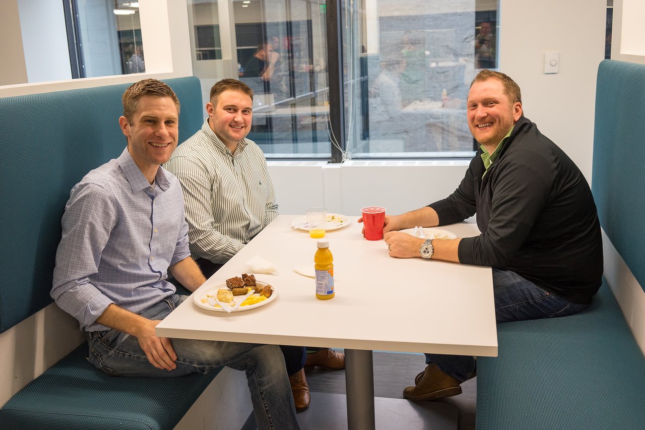 Three men sitting at a booth, smiling and having breakfast with drinks and plates of food.