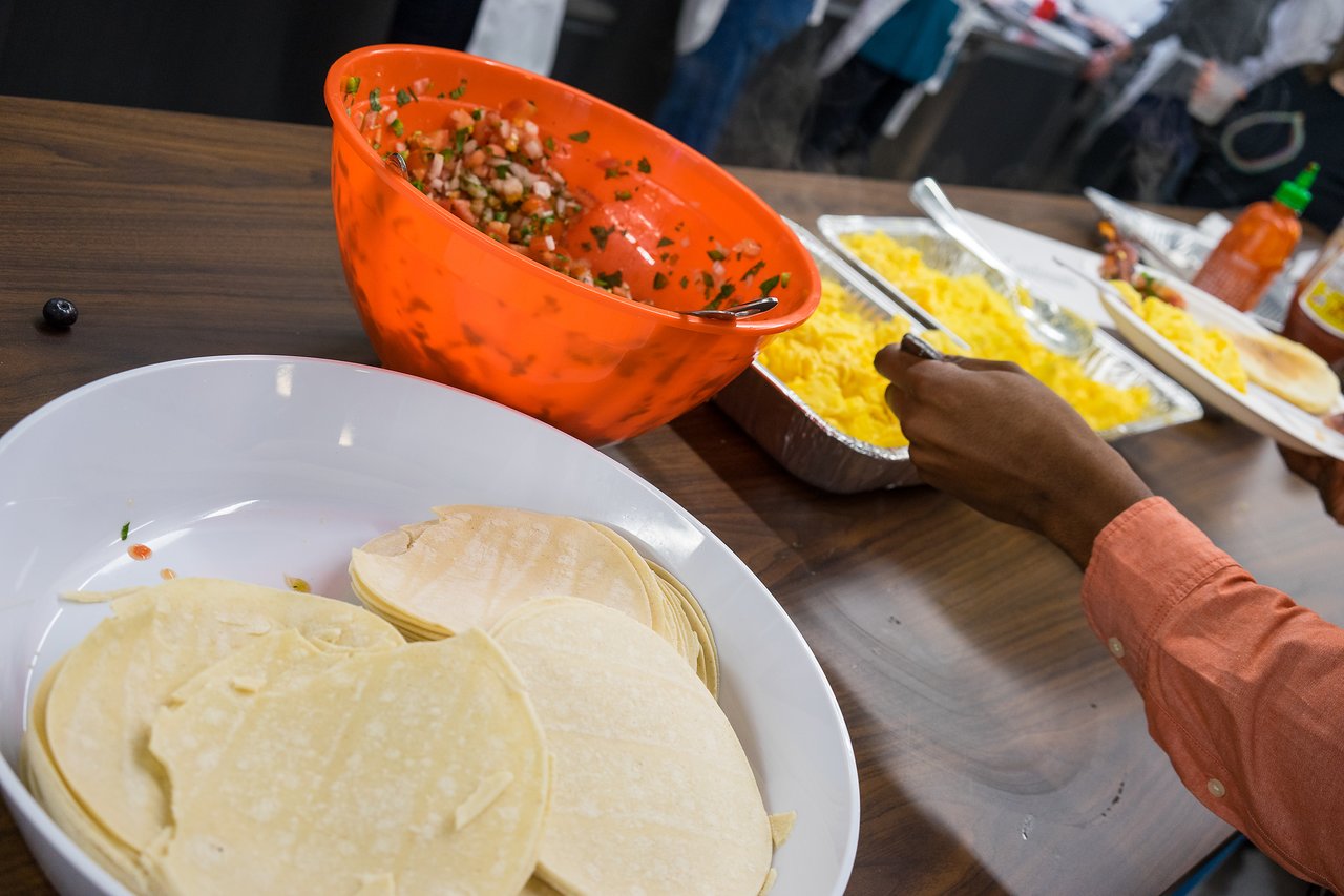 A person serves scrambled eggs onto their plate at a breakfast buffet with tortillas and salsa.