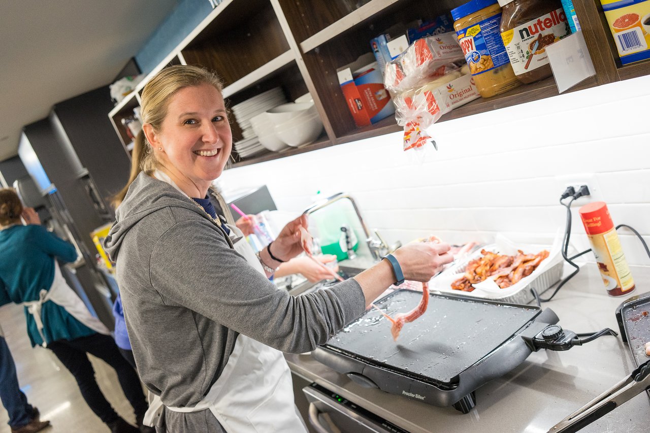 A woman wearing an apron smiles while cooking bacon on a griddle in a kitchen.