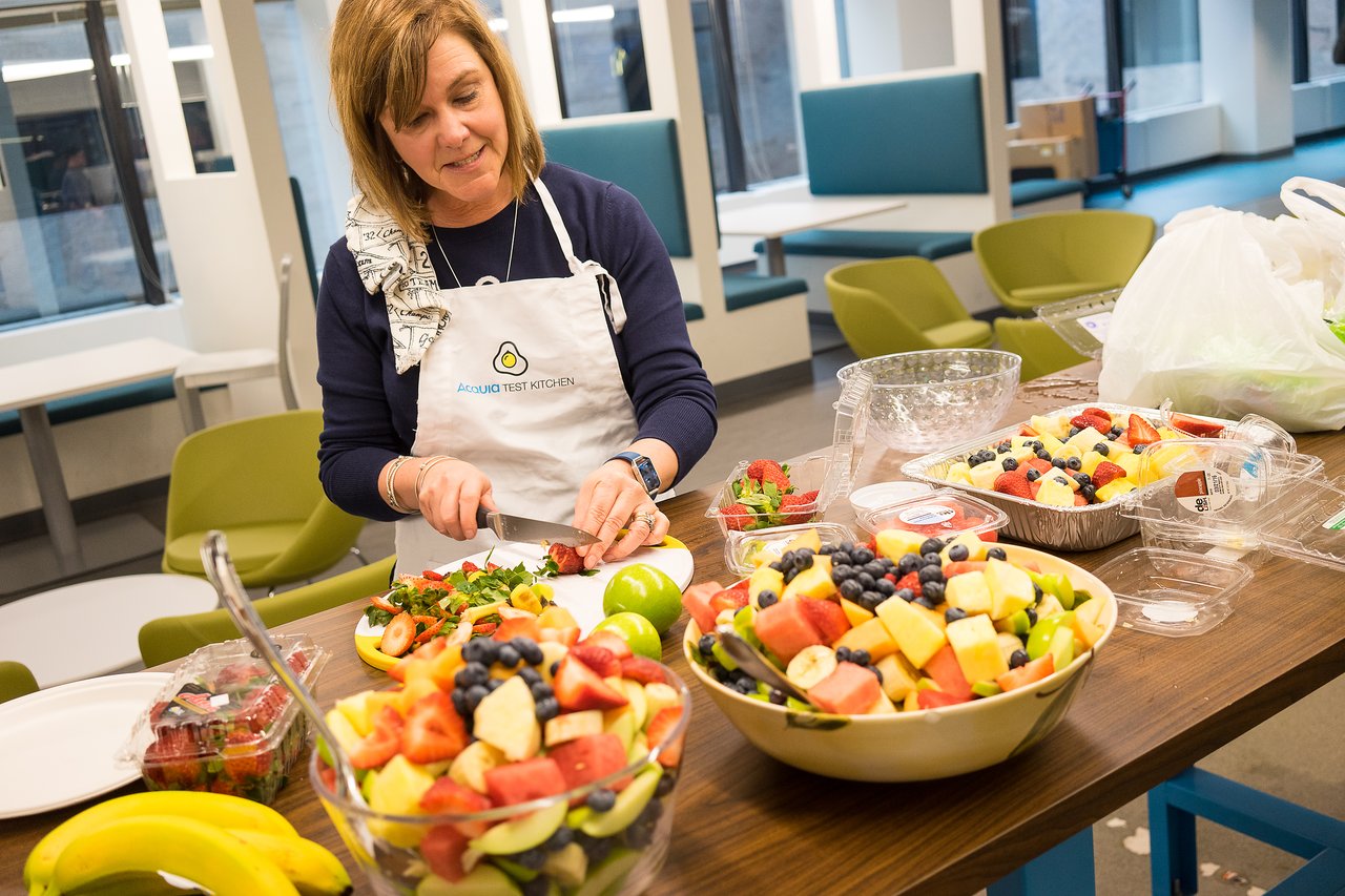 A woman wearing an "Acquia Test Kitchen" apron prepares fresh fruit salad at a table with various ingredients.