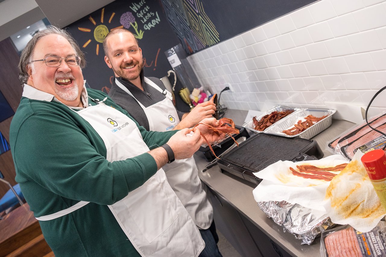 Two people wearing aprons smile while cooking bacon on a griddle in a kitchen.