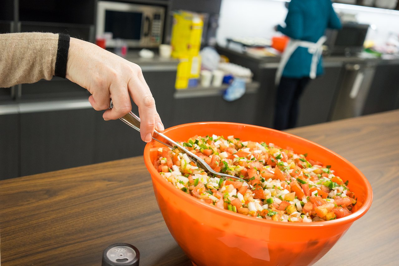 A person uses tongs to serve fresh salsa from a large orange bowl in a kitchen setting.