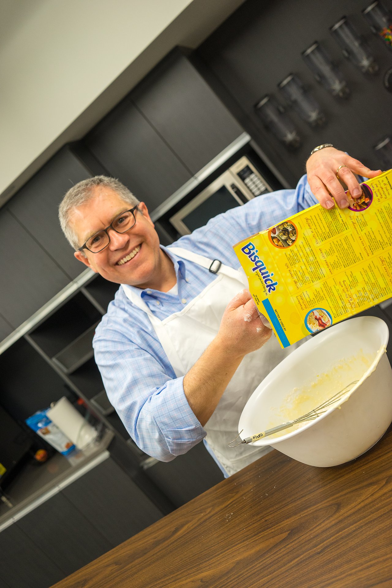 A smiling man in an apron holds a Bisquick box while preparing batter in a mixing bowl.