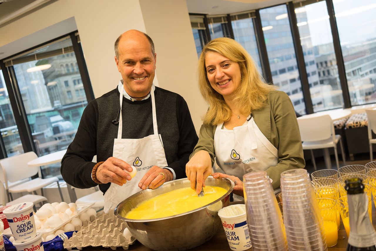 Two people wearing aprons smile while mixing eggs in a large bowl at a breakfast event.