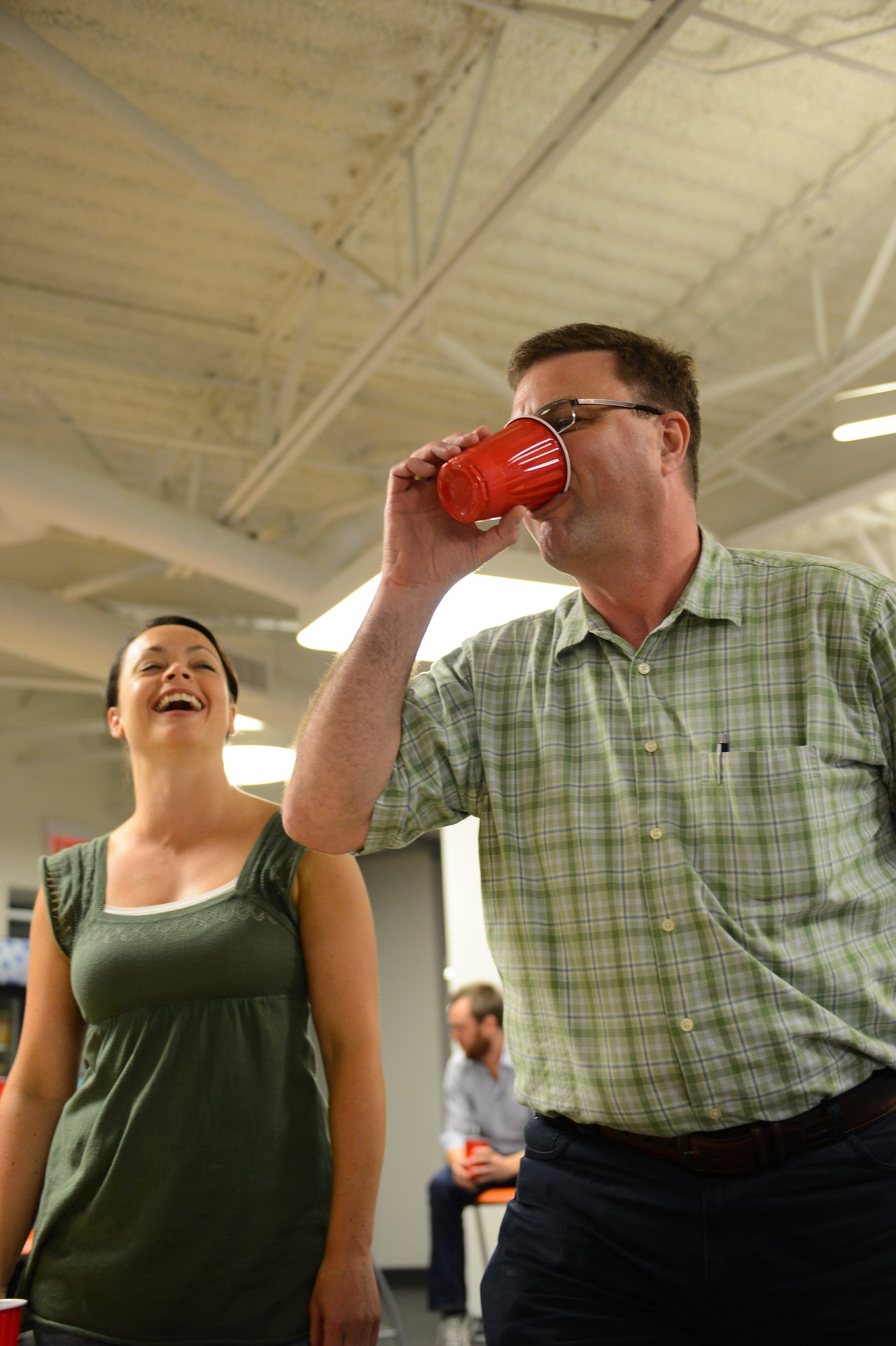 A man in a plaid shirt drinks from a red cup while a woman nearby laughs.