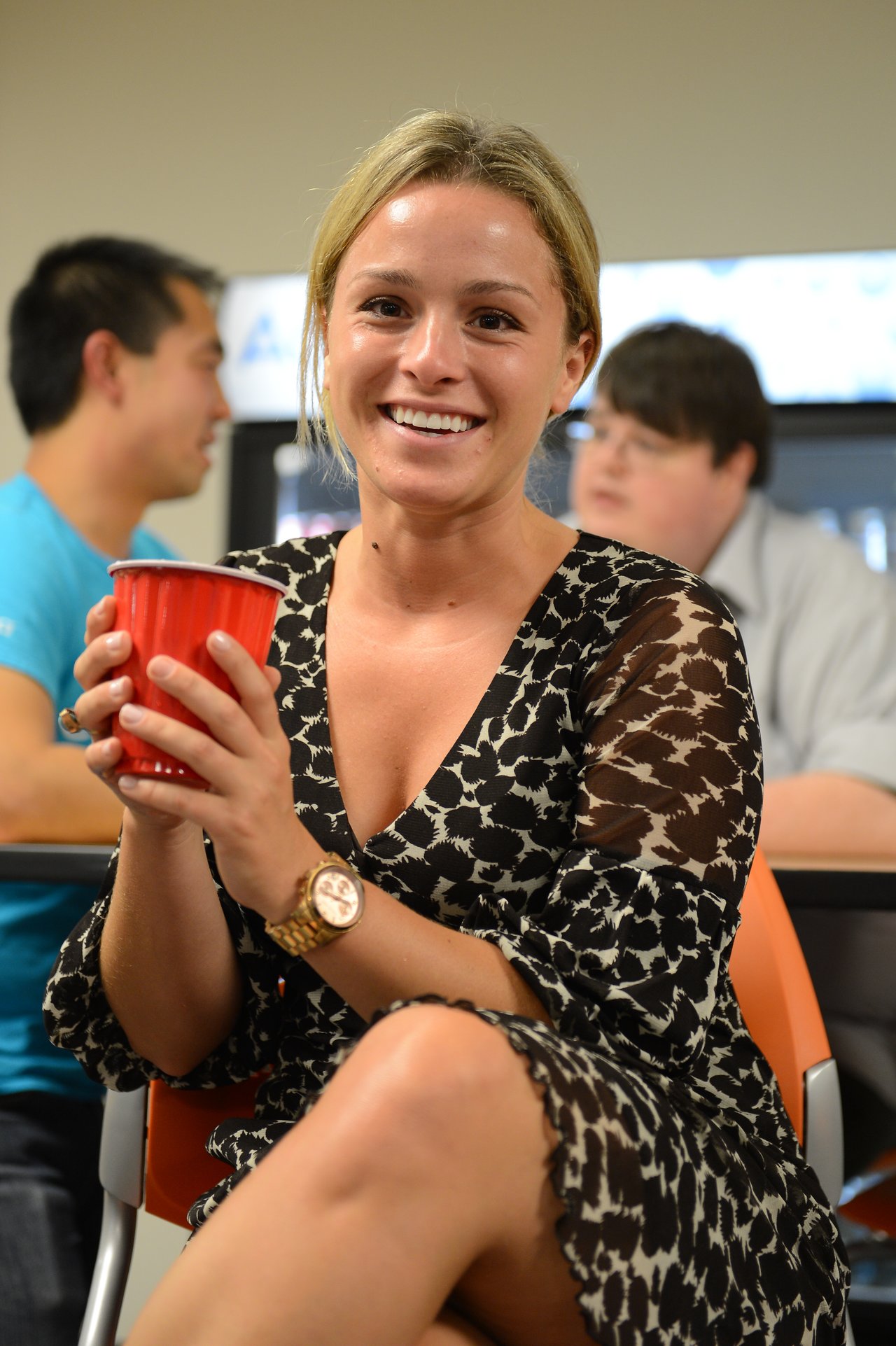 A smiling woman holds a red cup at a party, with people socializing in the background.