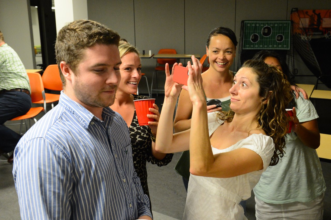 A woman playfully takes a close-up photo of a man while others smile and watch at a party.