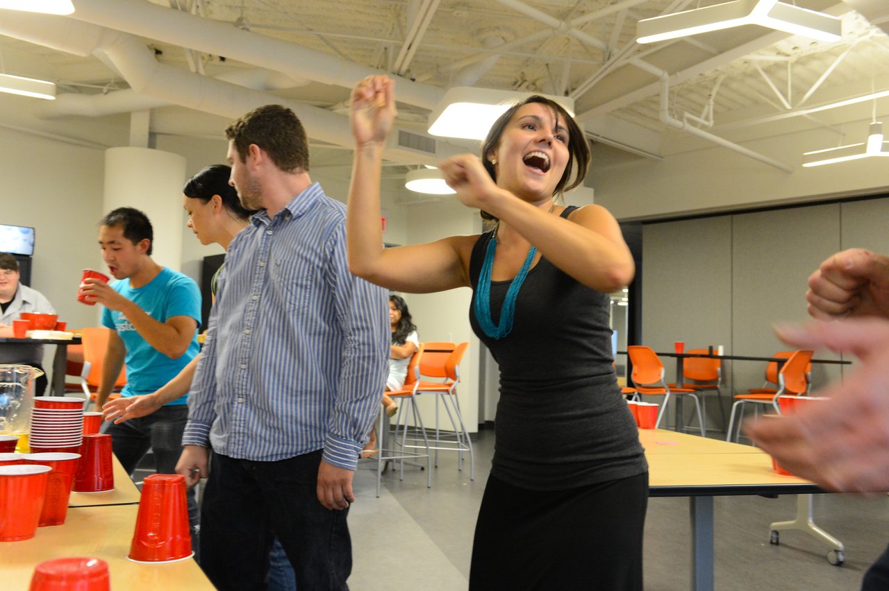 A woman in a black dress dances and smiles while others play a cup-stacking game at a party.