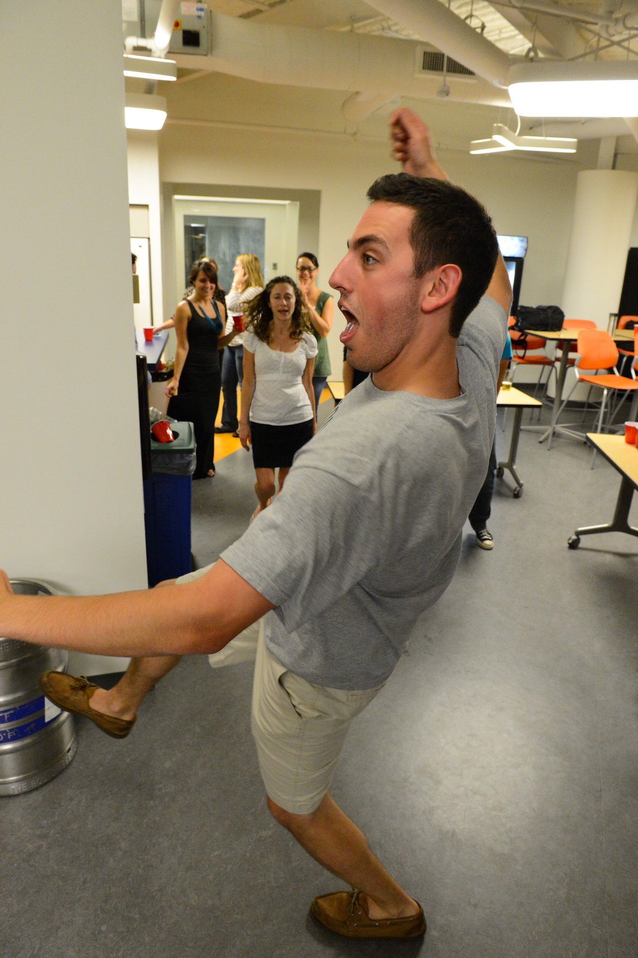 A man in a gray shirt energetically dances, while a group of people in the background watch and react.