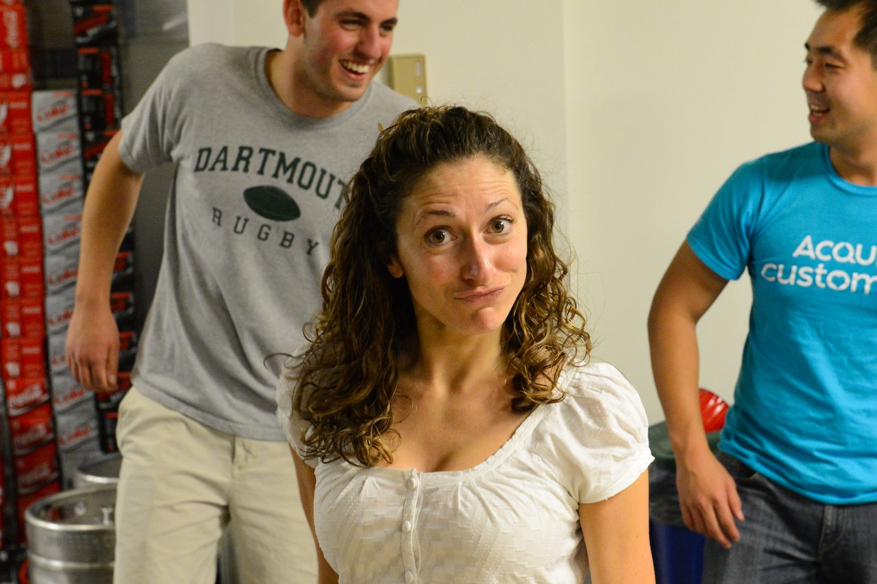 A woman in a white shirt makes a playful expression while two men behind her smile and laugh.
