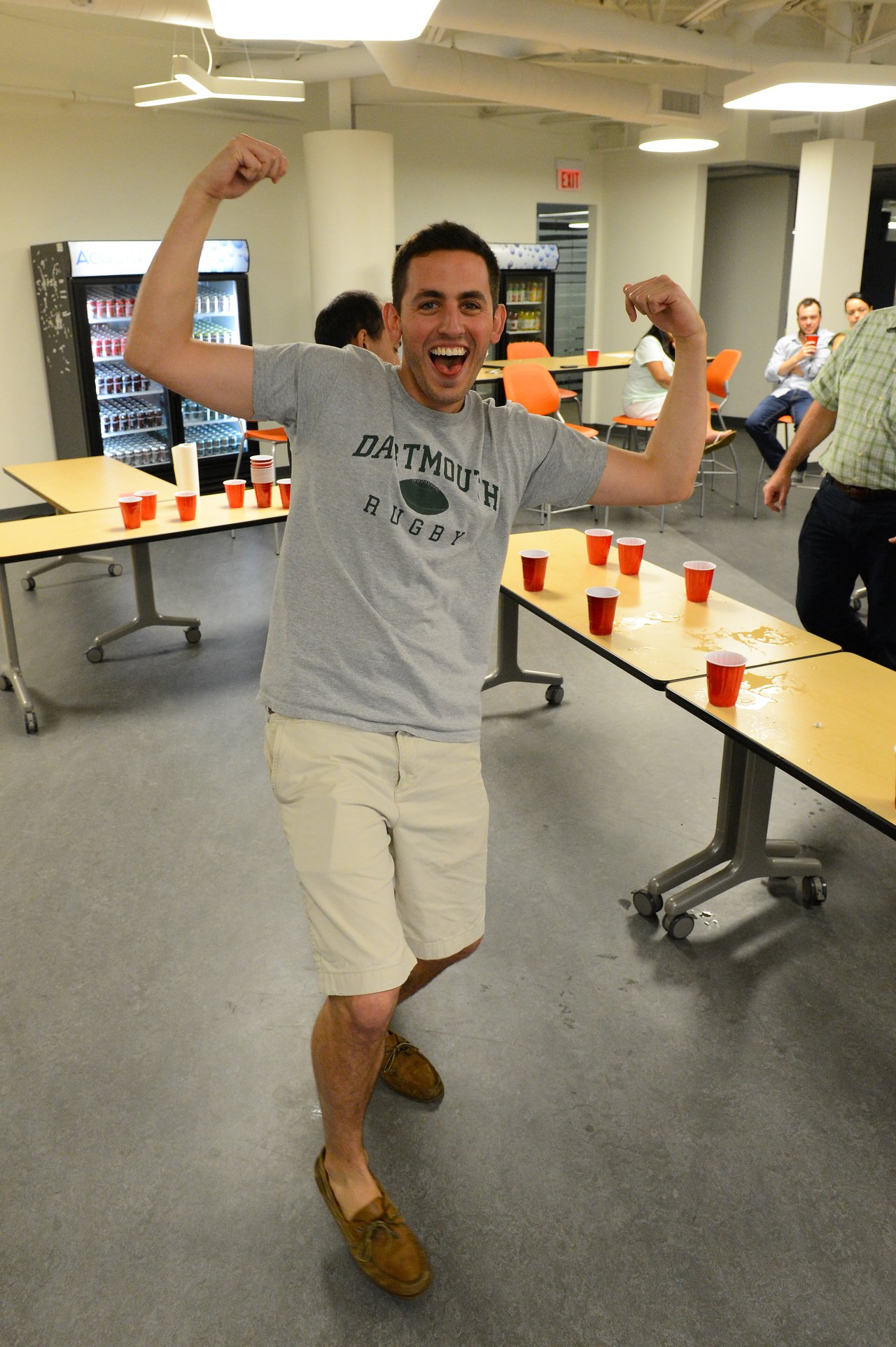 A man in a gray t-shirt flexes his arms and smiles at a party with red cups on tables.