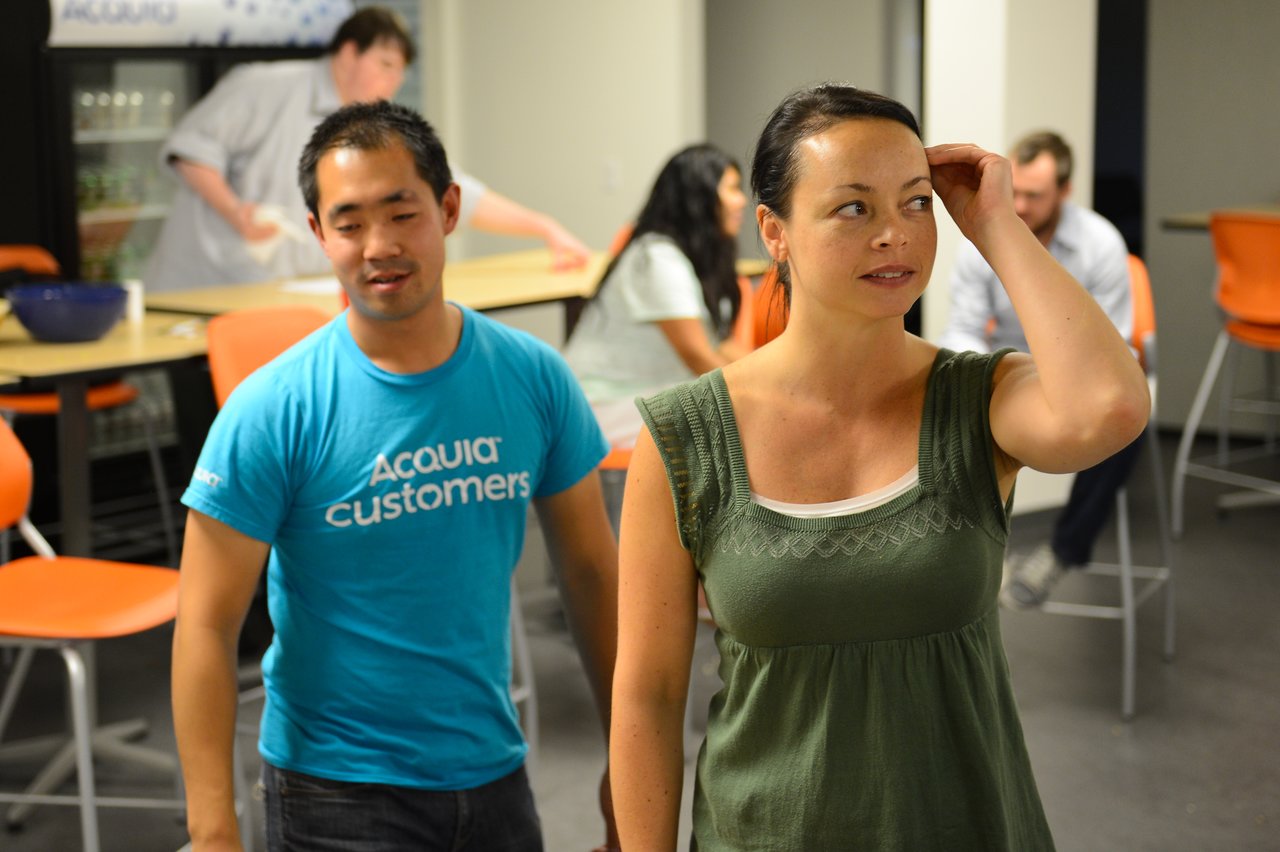 A man in a blue "Acquia Customers" shirt and a woman in green talk at a casual indoor gathering.