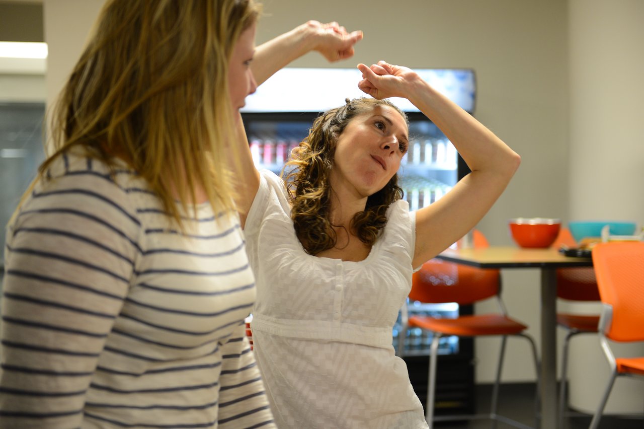 Two women at a party, one dancing with raised arms while the other watches.
