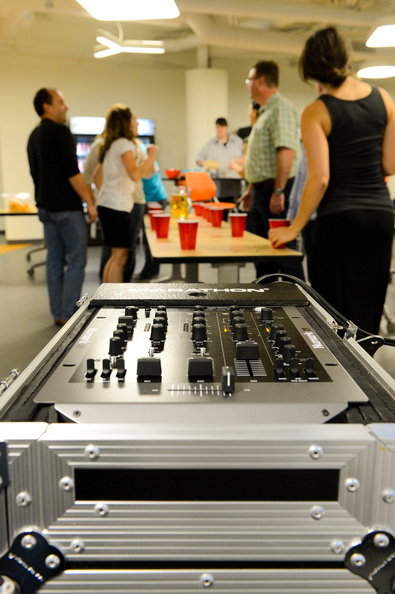 A DJ mixer in the foreground with people socializing and playing a party game with red cups in the background.