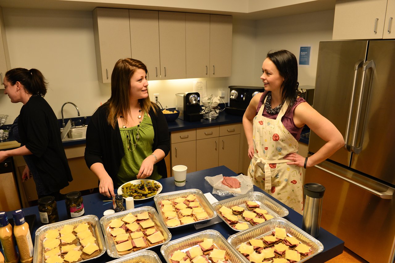 Two women stand in a kitchen, smiling and talking while preparing trays of sandwiches for a gathering.