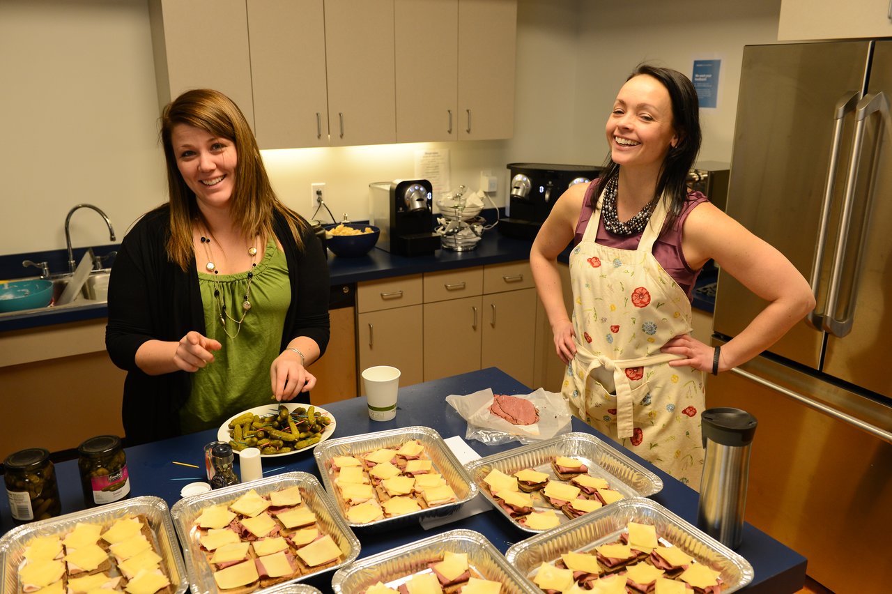 Two women in a kitchen preparing food, with trays of sandwiches and a bowl of pickles on the counter.
