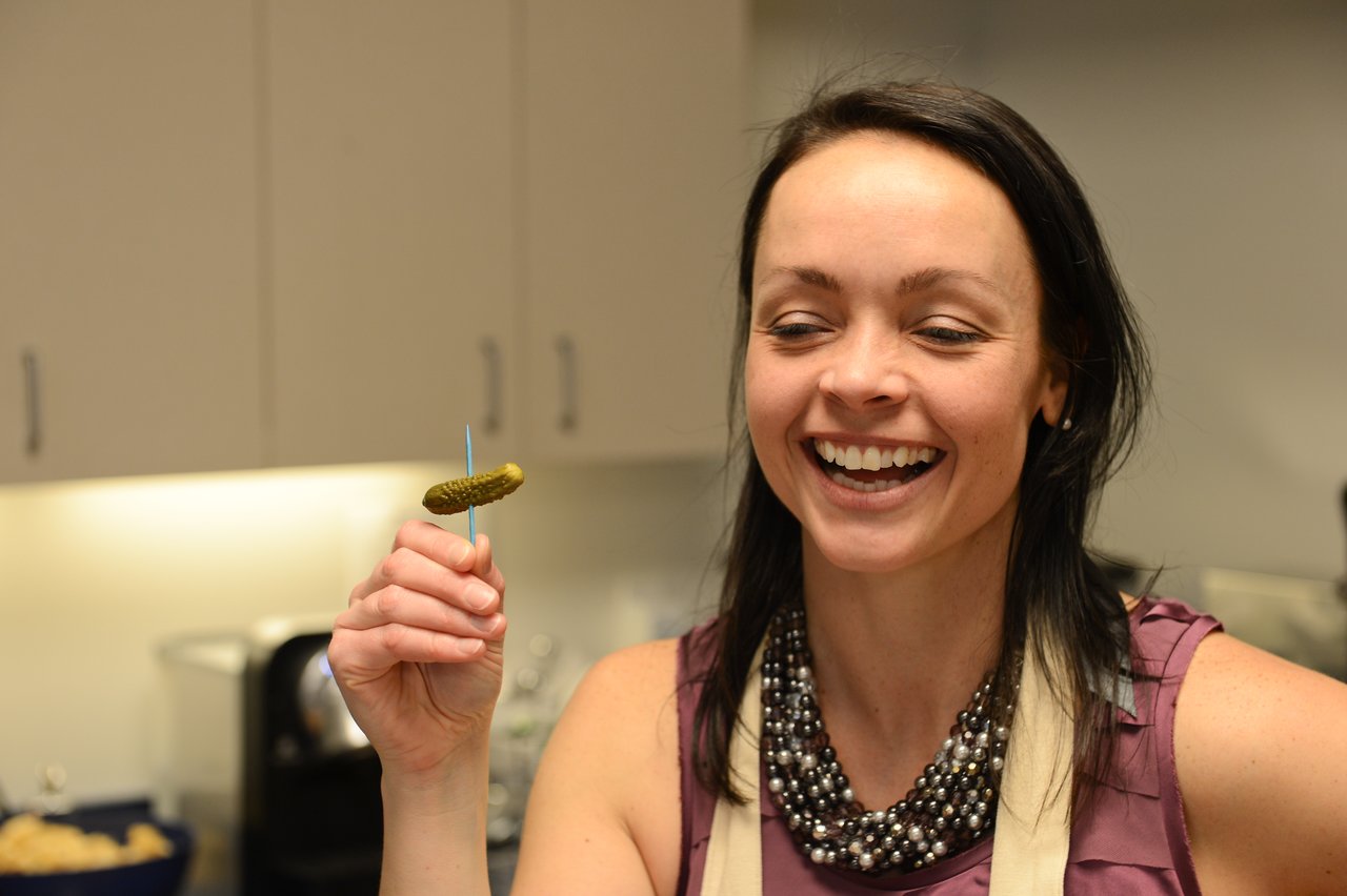 A smiling woman holds a small pickle on a toothpick at a party in a kitchen setting.